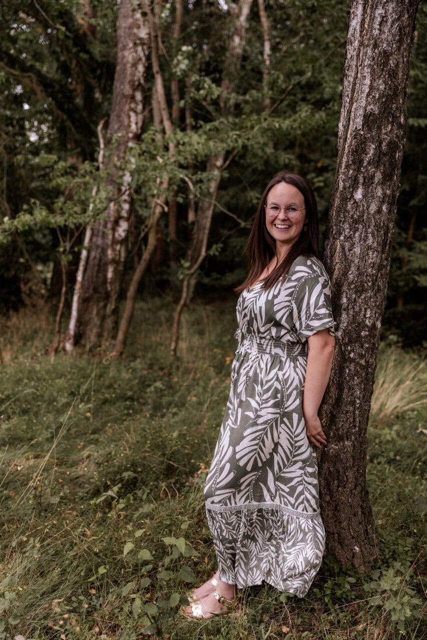 Frau in Waldkleid lehnt an Baum im Grünen Waldgebiet.
